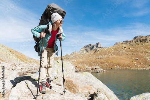 A hiker girl in sunglasses with a backpack and tracking sticks rises to a high rock against the background of rocks and a high mountain lake