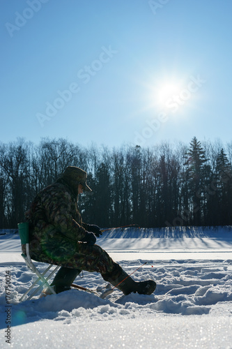 One euriopean man is fishing close to Ice hole at winter sunny day under blue sky photo