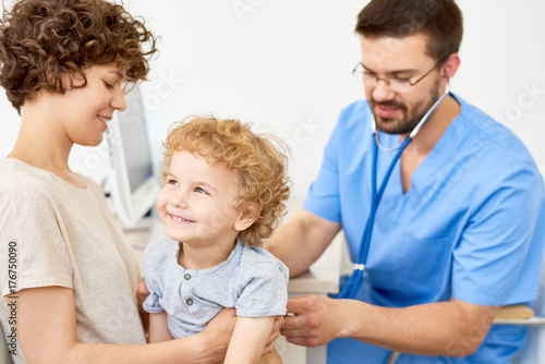 Portrait of happy little boy  sitting on mothers lap in doctors office while pediatrician listening to breathing using stethoscope