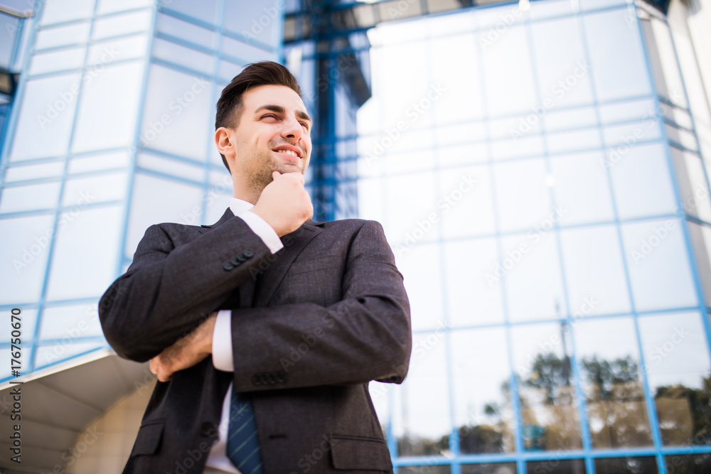 Confident thoughtful businessman in a suit holding hand on chin and looking away while standing outdoors with office building in the background Confident thoughtful businessman in a suit holding hand on chin and looking away while standing outdoors with office building in the background