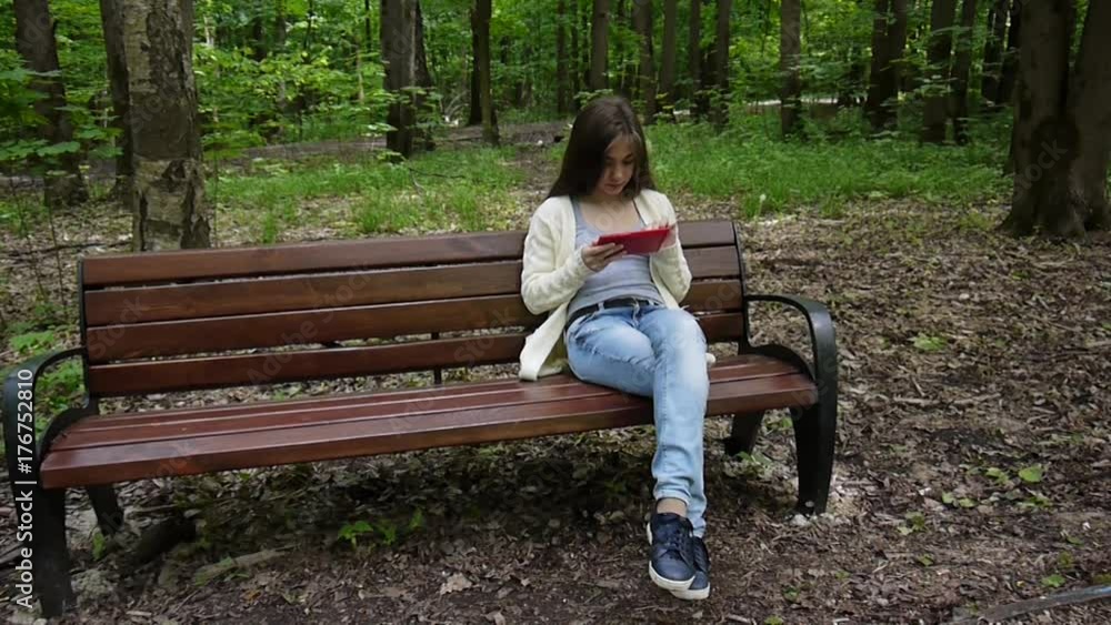 Beautiful teenager girl with tablet computer sits on the bench in Park. Movie of static camera.