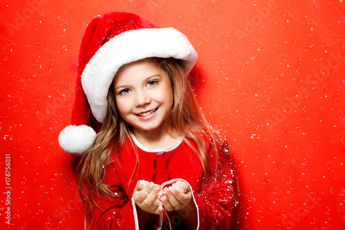 Happy smiling girl in Christmas cap under the snow on red background. Christmas.