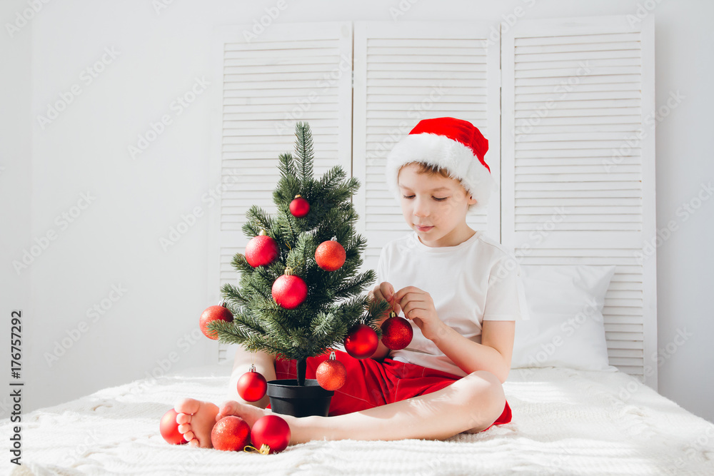 Boy in red Santa hat decorates a small Christmas tree balls Boy in red Santa hat decorates a small Christmas tree balls