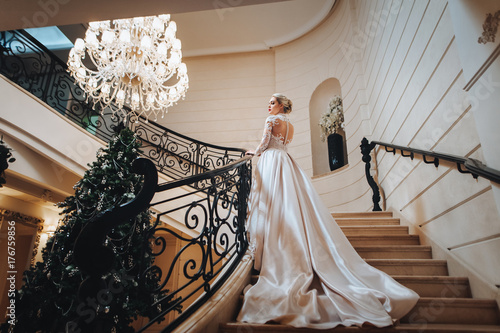 Beautiful bride in a white dress with long train is climbs up the stairs in a classic interior. 