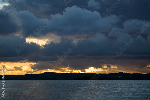 View of dark and dramatic clouds, rays of light and a lake in Finland at sunset in the summer.