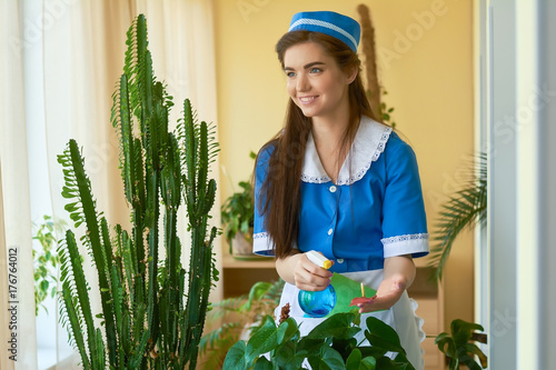 Smiling housemaid at work. Young woman water spraying plant.