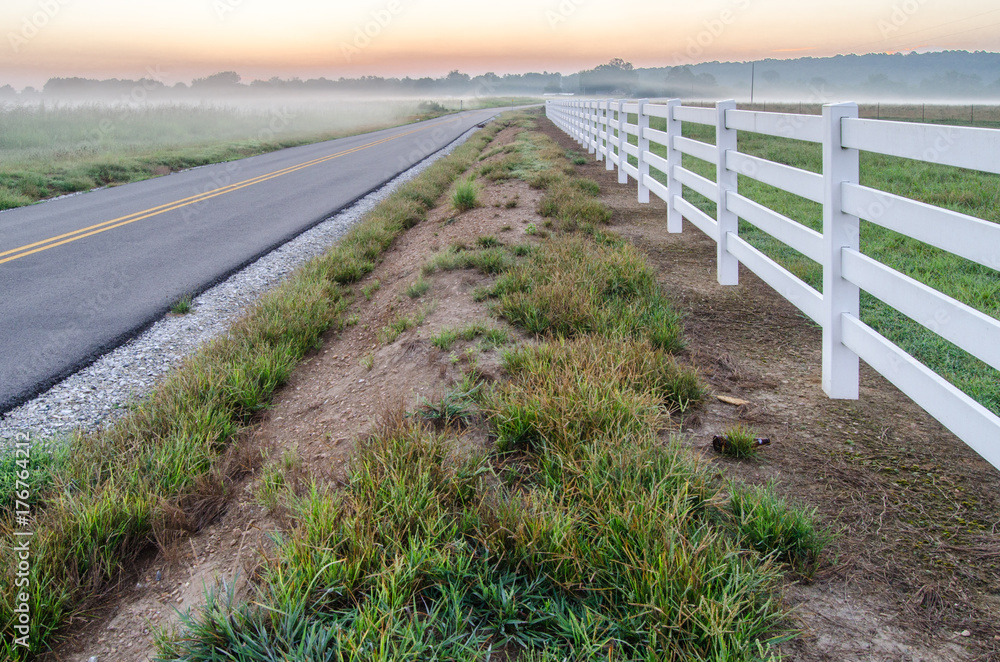 A white fence parallels a country road shortly before a foggy sunrise near Oxford, Alabama, USA