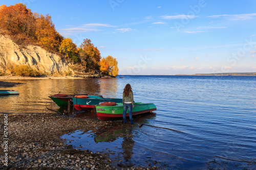 The girl stands at the moored boats to the stony shore, anchored on the shore against the backdrop of a forested slope, and looks out into the distance to the river and the sky. Autumn