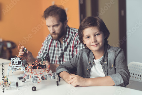 A man helps a boy with a robot assembly. The boy looks at the camera while the man assembles the robot