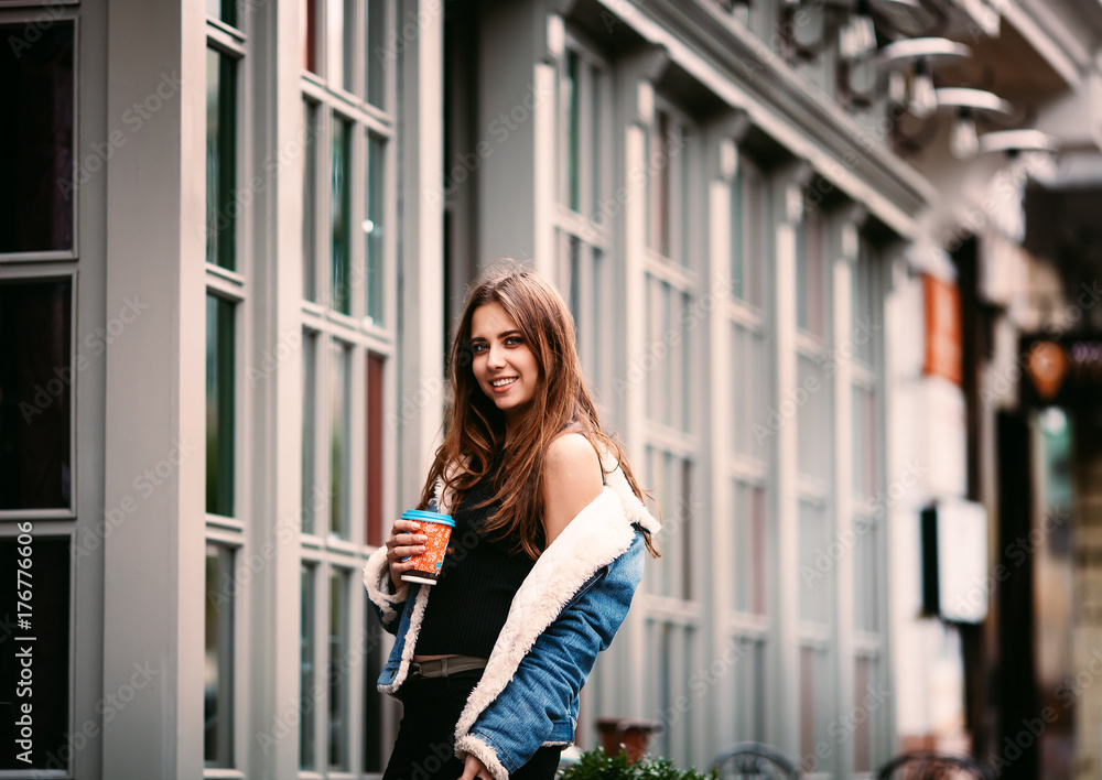 The concept of street fashion. portrait of Young girl dressed in a fashionable outfit. Posing against the window of the boutique girl smiling and drinking coffee
