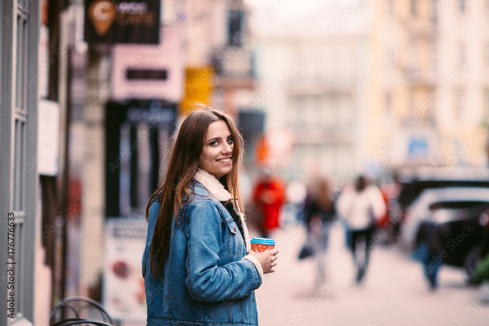 Outdoor portrait of young beautiful happy smiling girl posing on street. Model wearing stylish warm clothes.Keeps coffee.