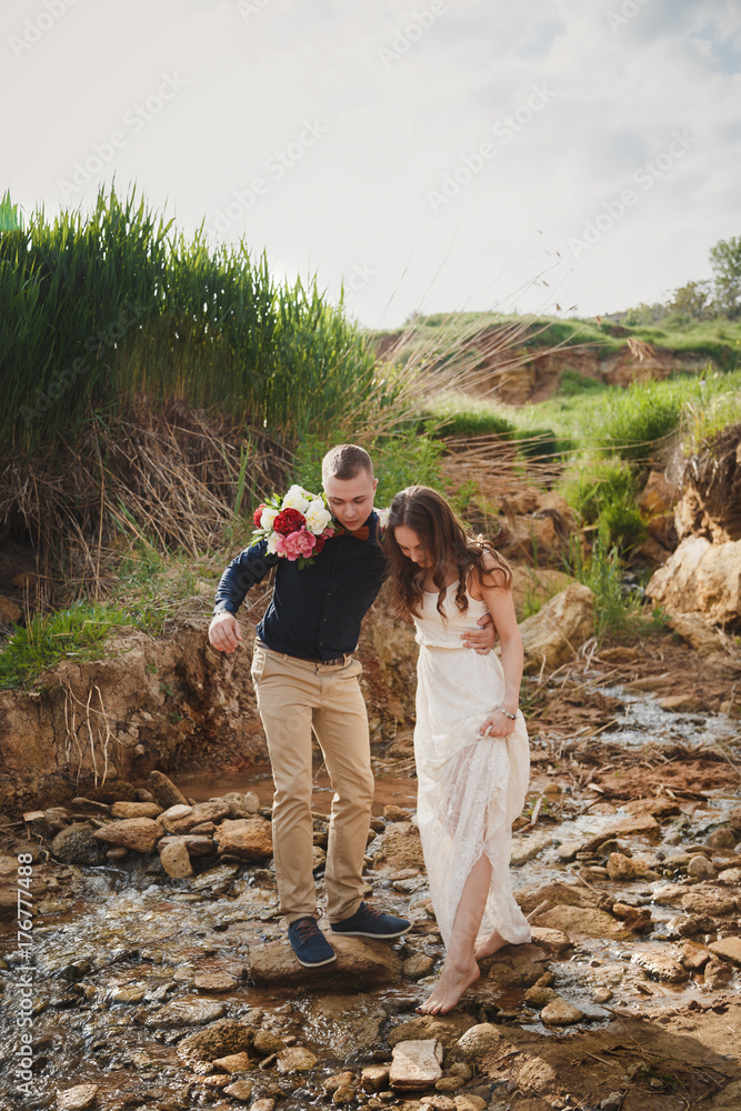 Outdoor beach wedding ceremony, stylish happy smiling groom and bride are crossing small river