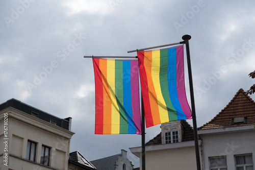 Two rainbow flags from Antwerp Pride hanging on flagpoles, bright against a gloomy sky. Small houses in the background. Gay flags from Antwerp Pride festival in the town centre near Grote Markt.