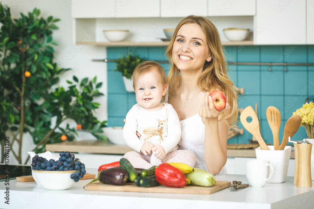 Young mother looking at camera and smiling, cooking and playing with her baby daughter in a modern kitchen setting. Healthy food concept.