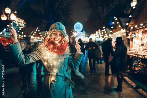 Outdoor photo of young beautiful happy smiling girl holding sparklers, posing in street. Festive Christmas fair on background. Model wearing stylish winter coat, knitted beanie hat, scarf.