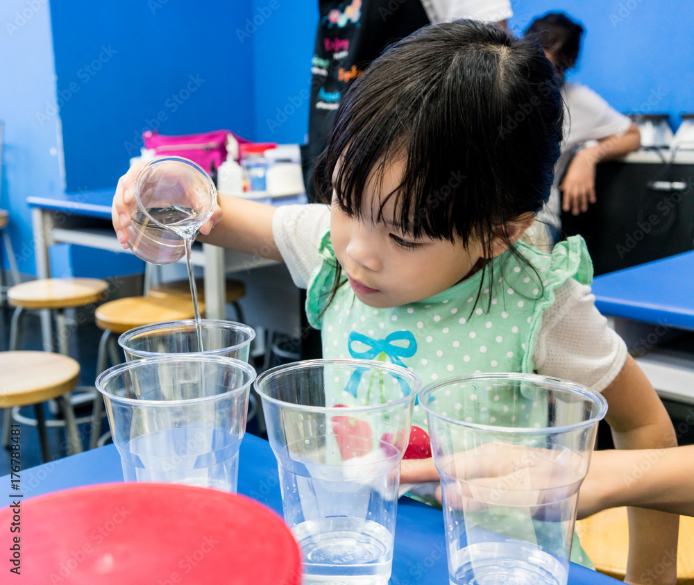 Kindergarten girl pouring clear solution into a cup on blue table. Kindergarten girl pouring clear solution into a cup on blue table.