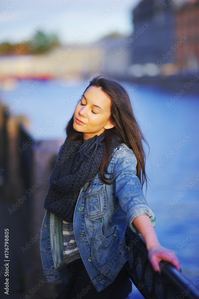Portrait of a stylish longhaired woman meditating on bridge with closed eyes in the sunset blurred background of urban buildings. Close-up
