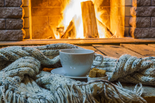 Cup of tea and sugar,  wool scarf near  cozy fireplace, in country house, winter vacation, horizontal.