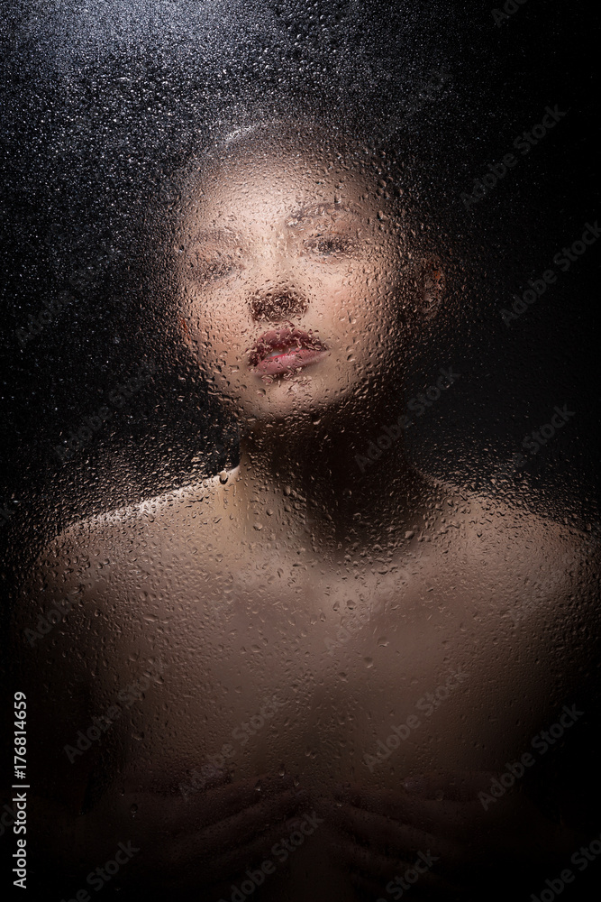 Portrait of a bald girl behind a wet glass on a black background.