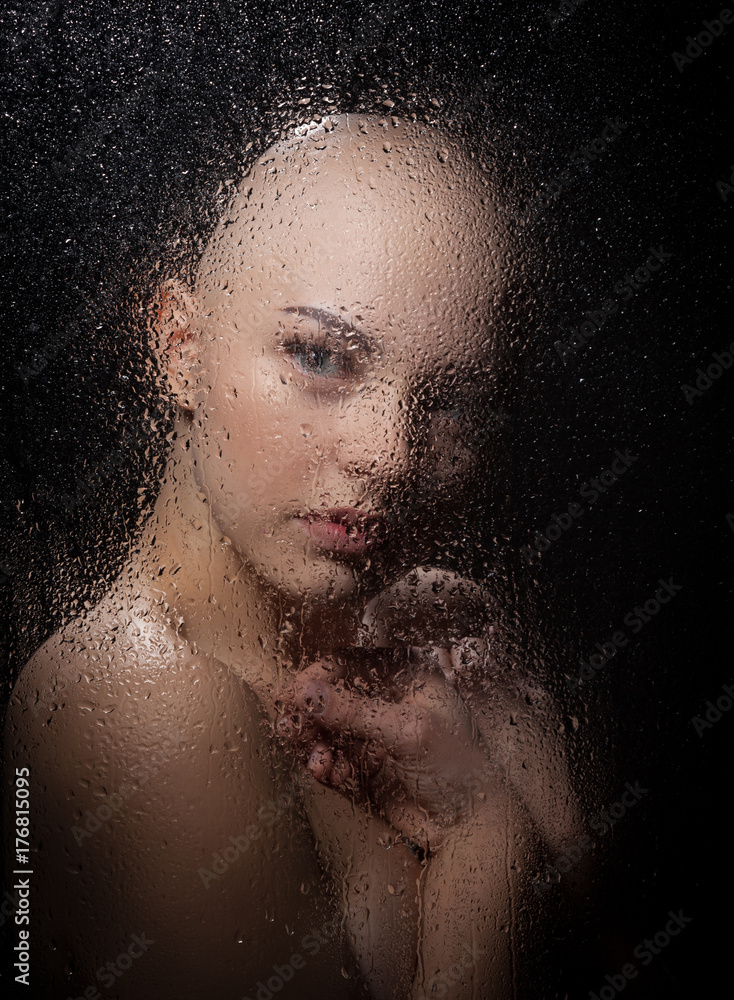 Portrait of a bald girl behind a wet glass on a black background.