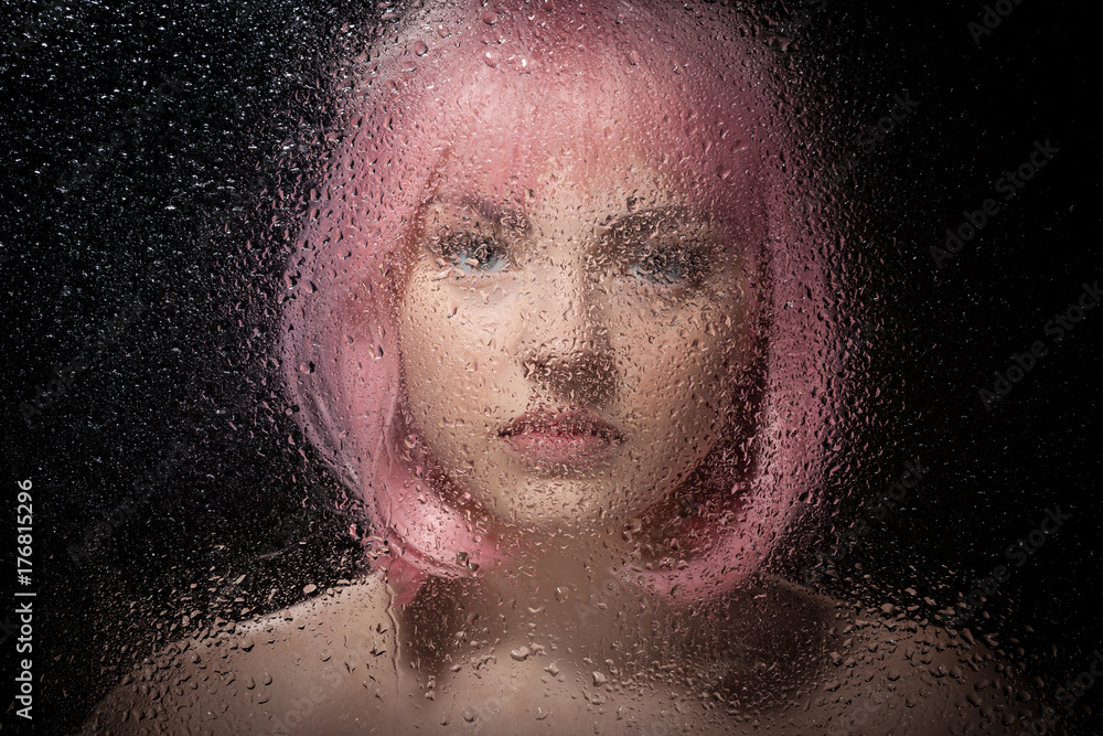 Portrait of a bald girl with pink hair behind a wet glass on a black background.
