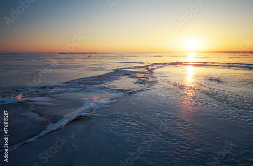 Winter landscape with cracks on the frozen lake near the shore at sunset