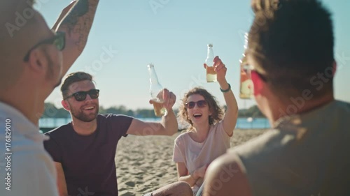 Young people clinking bottles, drinking and having fun on the beach. Medium shot. Soft Focus.