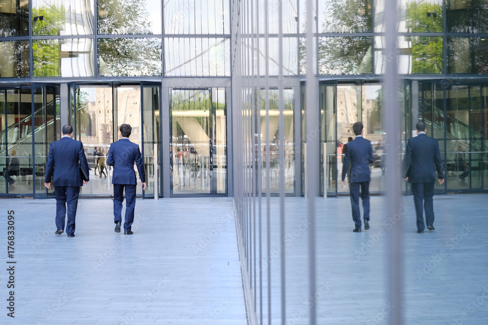 Two businessmen walk along the square to the business center, reflected in the glass of the facade