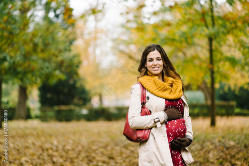 Happy pregnant woman with scarf and raincoat touching her belly in autumn. Brunette female wearing raincoat and warm yellow scarf.