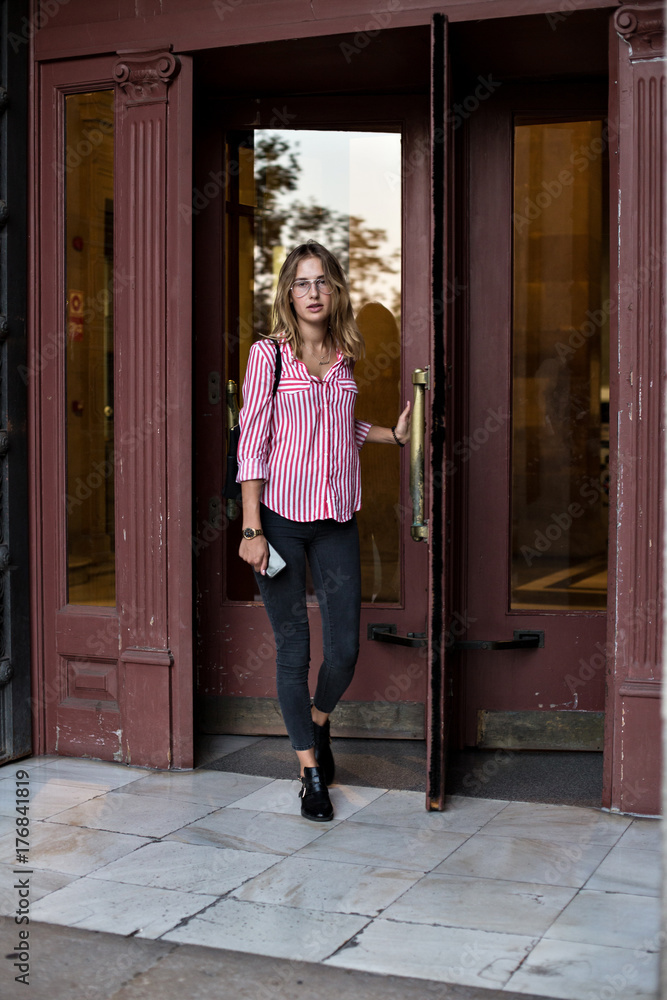 Pretty and cute feminine woman in skinny jeans and red shirt leaves building through vintage wooden rotating doors on warm summer evening, on way to business meeting Pretty and cute feminine woman in skinny jeans and red shirt leaves building through vintage wooden rotating doors on warm summer evening, on way to business meeting