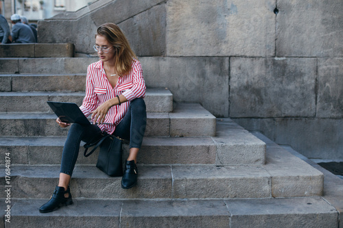 Fashionable trendy millennial hipster woman sits on stairs of university, works on laptop outdoors, freelance or out of office day, lunch break business woman always connected