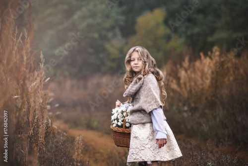 A beautiful little girl with a basket of wildflowers in her hand goes on a field road towards the forest.