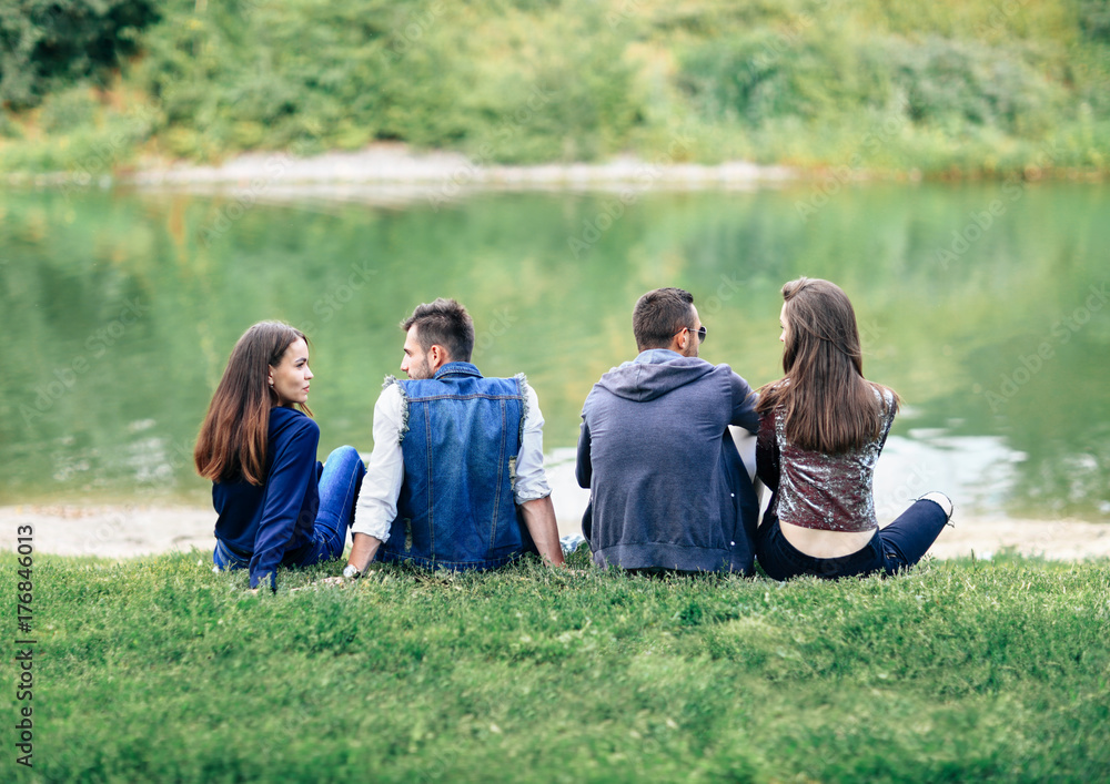 Obraz premium Friends sitting on grass by lake in park back view