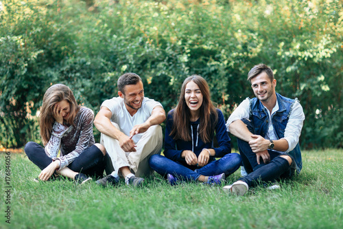 Schilderij op canvas Funny laughing friends sitting on grass with crossed legs
