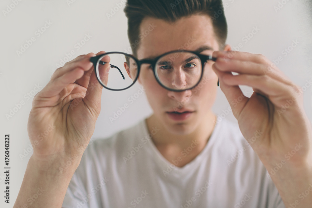 Closeup portrait of young man with glasses. He has eyesight problems and is squinting his eyes a little bit. Handsome guy is holding his eyeglasses right in front of camera with one hand. The concept Closeup portrait of young man with glasses. He has eyesight problems and is squinting his eyes a little bit. Handsome guy is holding his eyeglasses right in front of camera with one hand. The concept