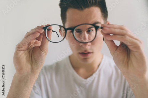 Closeup portrait of young man with glasses. He has eyesight problems and is squinting his eyes a little bit. Handsome guy is holding his eyeglasses right in front of camera with one hand. The concept photo