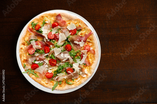 Prosciutto pizza with cherry tomatoes and rucola on dark brown table from above.