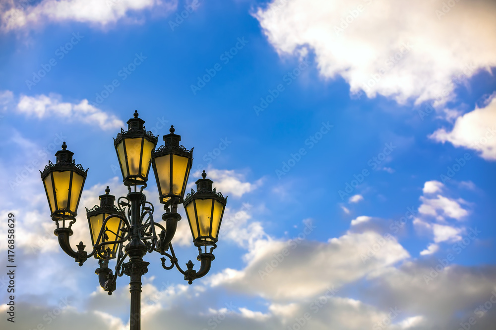 Street black lamppost Pushkin with a lamp made of cast iron against a background of a sunny blue sky with white clouds. City lamppost, Moscow, Russia. Street black lamppost Pushkin with a lamp made of cast iron against a background of a sunny blue sky with white clouds. City lamppost, Moscow, Russia.