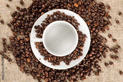 White coffee mug with plate among many of roasted coffee beans, top view