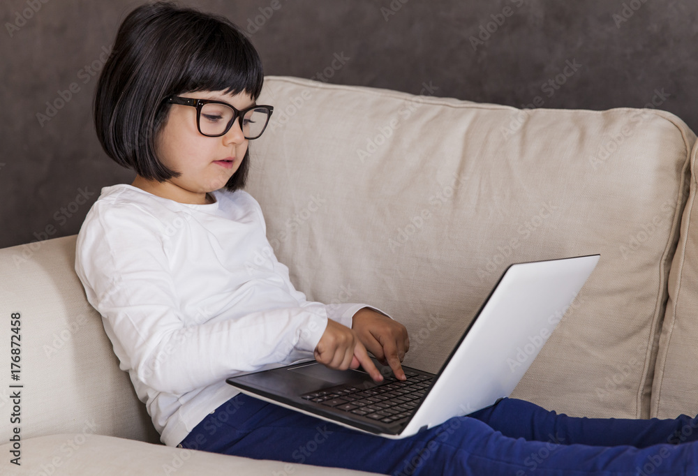Fototapeta premium Little girl with glasses using her laptop while sitting on the couch at home