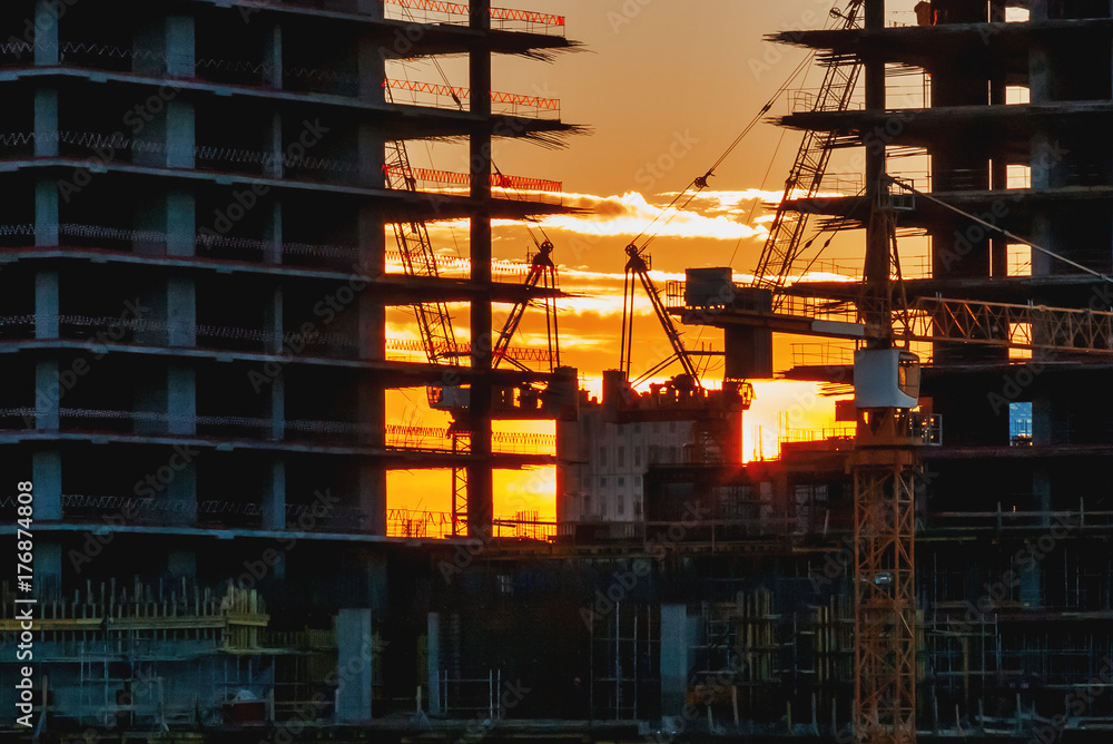 Sunset at the construction site. Silhouettes of cranes and unfinished floors on orange sky background. Moscow, Russia.