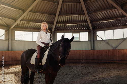 Beautiful elegant young blonde girl sits on a her black horse dressing uniform competition white blouse shirt and brown pants.