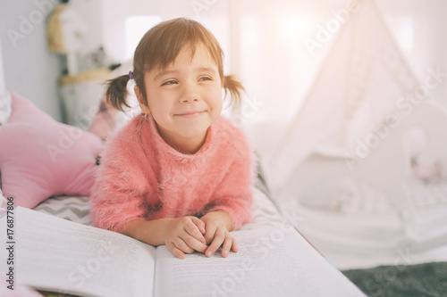 Cute little kid girl is reading a book at home. Funny lovely child having fun in kids room.