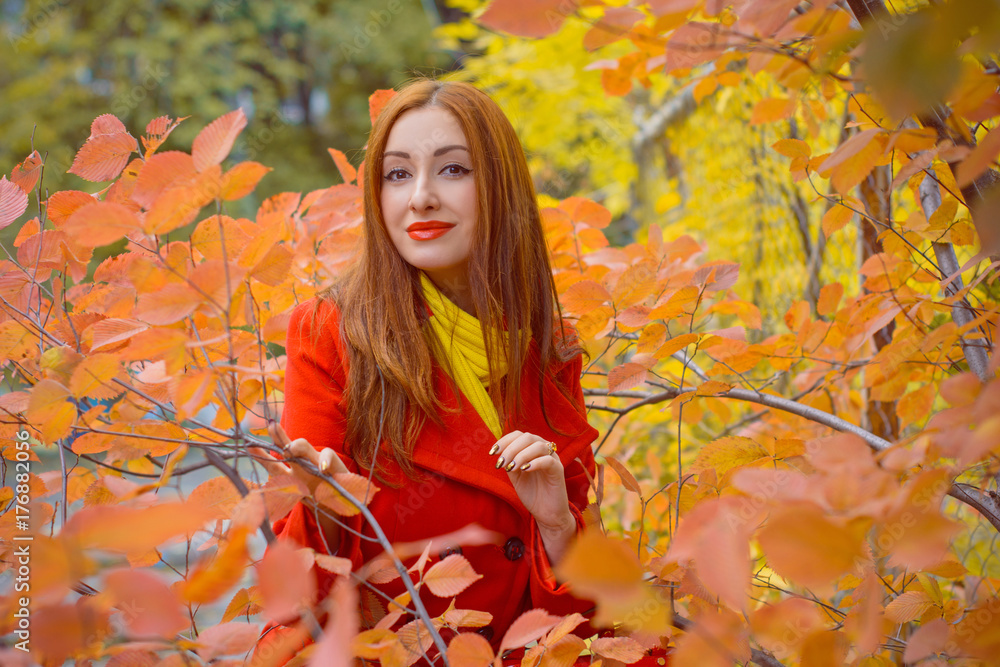 Cute and nice young beautiful woman in a red casual coat and red-yellow scarf walks in autumn city park. Pretty view of walking woman outside