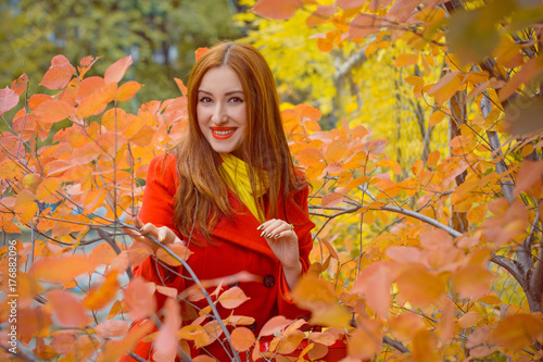 Cute and nice young beautiful woman in a red casual coat and red-yellow scarf walks in autumn city park. Pretty view of walking woman outside