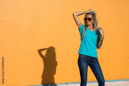 Beautiful blonde in sunglasses corrects luxurious hair. Portrait on the background of bright orange wall. Modern hipster girl with tattoos. photo