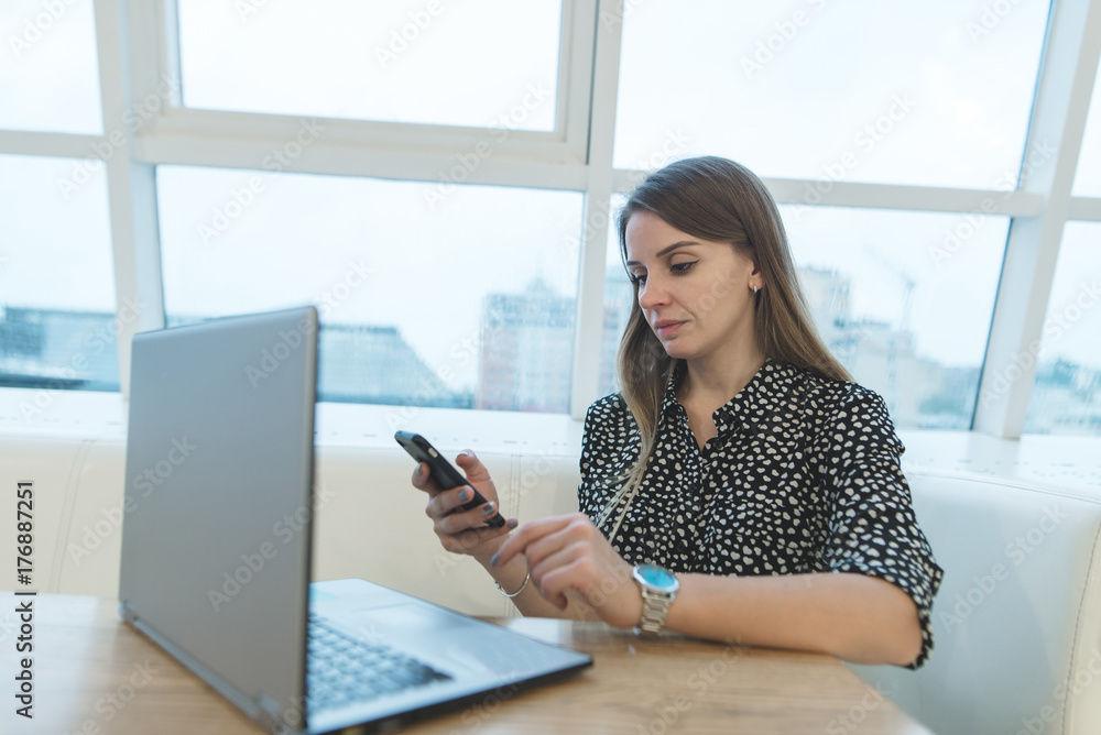 Beautiful business woman sitting in a cafe near a laptop and using a smartphone. A focused look at the phone. Modern woman. Lifestyle Beautiful business woman sitting in a cafe near a laptop and using a smartphone. A focused look at the phone. Modern woman. Lifestyle