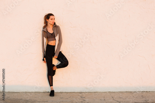 Young beautiful sport woman resting after running near white wall, looking at sunrise