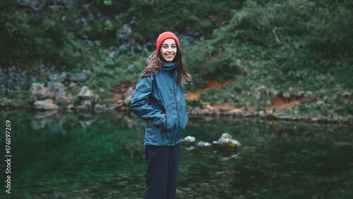 Fototapeta Naklejka Na Ścianę i Meble -  a woman tourist in waterproof clothes and red hat stands on the Bank of the mountain lake with turquoise water. Glanni waterfall, Iceland