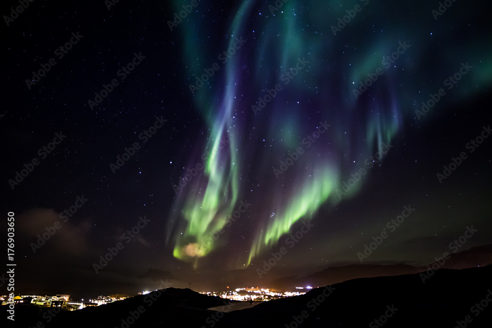 Glows of Northern Lights with shining stars on the sky over the mountains and highlighted city, Nuuk, Greenland